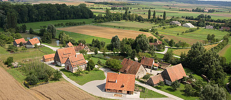 Fotoaufnahme eines Luftbildes der Baugruppe Ost. Die Häuser stehen in kleinen Gruppen und sind umgeben von Feldern, Wiesen und Bäumen. Im Hintergrund ist die weite Landschaft des Museums und der Umgebung erkennbar. 