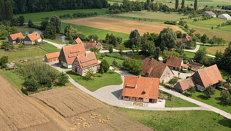 FFM_Baugruppe_Ost Fotoaufnahme eines Luftbildes der Baugruppe Ost. Die Häuser stehen in kleinen Gruppen und sind umgeben von Feldern, Wiesen und Bäumen. Im Hintergrund ist die weite Landschaft des Museums und der Umgebung erkennbar.