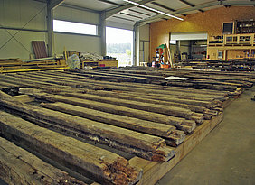 Die Balkenlage des Erdgeschosses in der Abbundhalle der Firma Lederer Fotoaufnahme der Abbundhalle der Firma Lederer. In dieser wurde die bauzeitliche Balkenlage des Erdgeschosses sortiert und der Reihe nach auf dem Boden ausgelegt. Einige Holzbalken sind beschädigt. Das Tor der Halle ist geöffnet, an den Wänden befinden sich Regale und Baumaterialien.