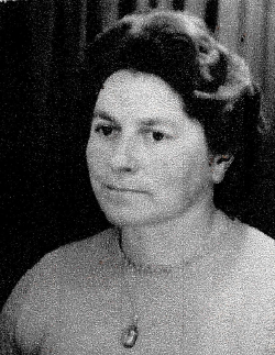 Das Bild zeigt eine Frau mit dunklen Haaren. Sie steht seitlich zur Kamera. Sie trägt einen Pullover und eine Kette.