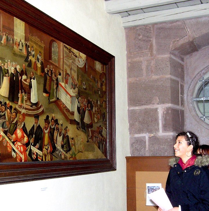 FFM_Wozu_wir_uns_bekennen Fotoaufnahme der Führung „Bekenntnis – wozu wir uns bekennen“ in der Bad Windsheimer Spitalkirche. An einer Wand hängt ein Gemälde im Holzrahmen. Davor stehen zwei Jugendliche, die es betrachten. Die Wand rechts daneben besteht aus Sandsteinquadern.