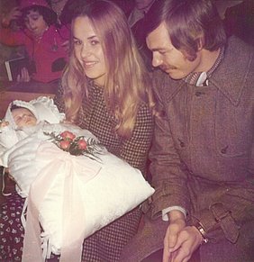 Eine Mutter hält das Taufkissen. Im Taufkissen ist ein Baby. Auf dem Taufkissen liegen Blüten. Neben der Mutter sitzt der Vater.