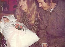 Eine Mutter hält das Taufkissen. Im Taufkissen ist ein Baby. Auf dem Taufkissen liegen Blüten. Neben der Mutter sitzt der Vater. 