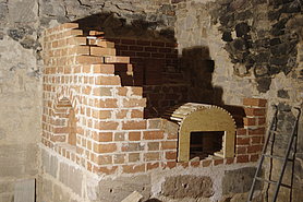 Fotoaufnahme des Schwitzofens in der Ecke. Der untere Teil des Ofens besteht aus Sandsteinquadern, der obere Teil ist etwa zur Hälfte mit Ziegelsteinen rekonstruiert worden, die andere Hälfte fehlt noch. Er weist an beiden Seite je eine rundbogige Öffnung auf. 