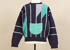 Der Pullover ist dunbkelblau. Er hat blaugraue Streifen. Zwei Stellen sind türkis. Vorne ist eine türkise Tasche aufgenäht.