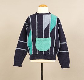 Der Pullover ist dunbkelblau. Er hat blaugraue Streifen. Zwei Stellen sind türkis. Vorne ist eine türkise Tasche aufgenäht.