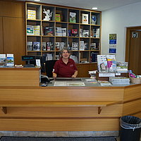 FFM_Kassenraum Fotoaufnahme des Kassenraumes im Korbhaus aus Knittelsbach am aktuellen Standort. Hinter dem Verkaufstresen steht eine Dame von Kassendienst. Auf dem Tisch und im Regal dahinter liegen verschiedene Flyer und andere Angebote des Museums aus.