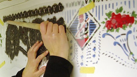 FFM_Schablone Fotoaufnahme einer Schablone, die mit Klebeband an einer Wand befestigt wurde. Das Muster der Schablone wird dabei mit Farbe und Pinsel stupfend auf die Wand gebracht. An der Wand befinden sich bereits Muster in verschiedenen Farben.