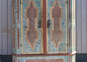 Der Schrank hat zwei Türen. Er steht auf einem Sockel. Er ist bunt bemalt. Die Bemalung ist in Felder aufgeteilt. Sie zeigt auch Blüten und Pflanzen.