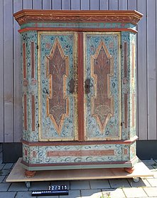 Der Schrank hat zwei Türen. Er steht auf einem Sockel. Er ist bunt bemalt. Die Bemalung ist in Felder aufgeteilt. Sie zeigt auch Blüten und Pflanzen.