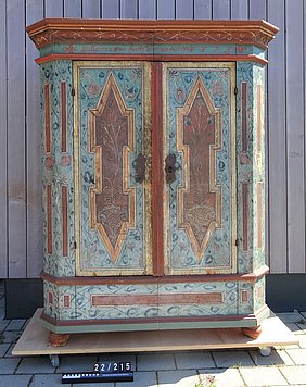 Der Schrank hat zwei Türen. Er steht auf einem Sockel. Er ist bunt bemalt. Die Bemalung ist in Felder aufgeteilt. Sie zeigt auch Blüten und Pflanzen.
