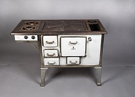 Der Herd ist weiß. Er hat sechs Herdplatten. Zwei Herdplatten werden mit Gas betrieben. Vier Herdplatten werden mit Holz betrieben. Er hat auch ein Wasserschiff, in dem man Wasser kochen kann.