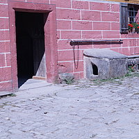 FFM_Muehle-Haupteingang Fotoaufnahme des Eingangsbereichs der Mühle aus Unterschlauersbach am aktuellen Standort. An den, mit Fensterläden versehenen, Fenstern stehen blühende Blumentöpfe. An der Hauswand lehnt u.a. ein Mahlstein.
