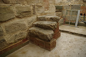 Stufen zur Schürkammer Fotoaufnahme der drei Sandsteinstufen, die vom Flur in die tieferliegende Schürkammer führen. Die Stufen befinden sich an der Mauer aus Sandsteinquadern und wurden mit Ziegelsteinen untermauert. Im Hintergrund ist eine weitere Mauer erkennbar, aus der zwei Rohre führen.