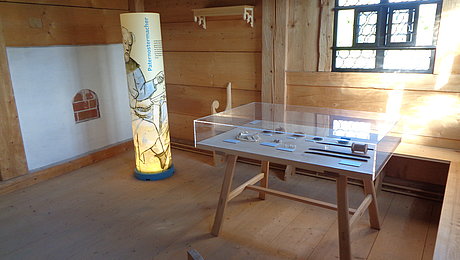 FFM_Badhaus_Wendelstein_Paternosterer_als_Mieter ein Tisch mit Exponaten, durch Glas geschützt sowie ein stehender Zylinder mit Ausstellungstext