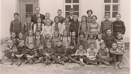 Historische schwarz-weiße Fotoaufnahme einer Schulklasse vor einem Haus. Die Klasse hat sich zum Klassenfoto in vier Reihen aufgestellt, die vorderen beiden Reihen sitzen bzw. knien. Der Lehrer steht links in der letzten Reihe. 