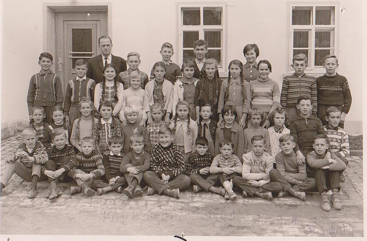 FFM_Fotos_Minuth Historische schwarz-weiße Fotoaufnahme einer Schulklasse vor einem Haus. Die Klasse hat sich zum Klassenfoto in vier Reihen aufgestellt, die vorderen beiden Reihen sitzen bzw. knien. Der Lehrer steht links in der letzten Reihe.