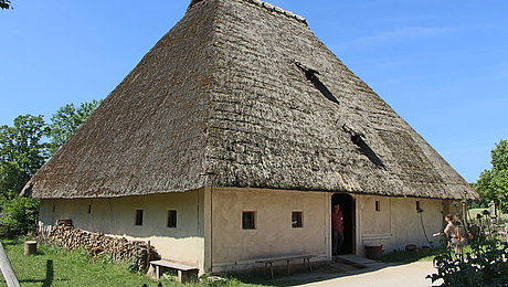 FFM_Hoefstetten Fotoaufnahme des Bauernhauses aus Höfstetten am aktuellen Standort. Das Haus hat einen quadratischen Grundriss und ein Strohdach. In der Tür steht ein Besucher, eine Frau geht auf ihn zu. An den Hauswänden stehen Holzbänke, an einer wurde Brennholz getrampelt.
