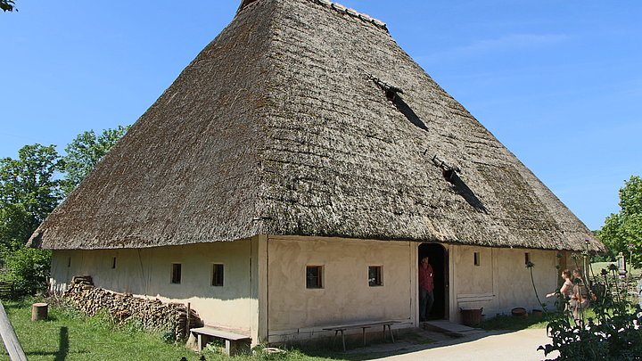 Fotoaufnahme des Bauernhauses aus Höfstetten am aktuellen Standort. Das Haus hat einen quadratischen Grundriss und ein Strohdach. In der Tür steht ein Besucher, eine Frau geht auf ihn zu. An den Hauswänden stehen Holzbänke, an einer wurde Brennholz getrampelt.  