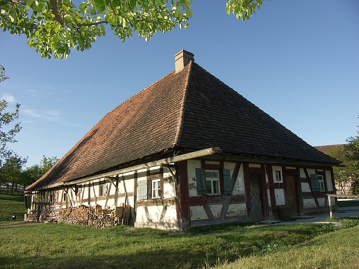 FFM_Schaeferei Fotoaufnahme der Schäferei aus Hambühl am aktuellen Standort. Das kleine Fachwerk wird von der Sonne beschienen. An einer Wand befinden sich eine lange hölzerne Dachrinne und ein Stapel Brennholz. Links oben ist das Geäst eines Baumes erkennbar.