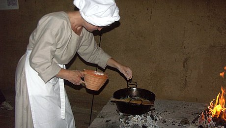 FFM_Kochen_MA Fotoaufnahme einer Frau in mittelalterlichem Gewand mit Schürze und Haube. Vor ihr steht ein Metalltopf über heißen Kohlen, in dem sie etwas zubereitet. Daneben brennt ein Feuer. Auf dem Herd steht außerdem eine kleine Kanne.