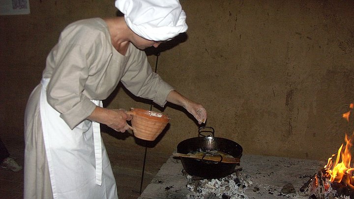 FFM_Kochen_MA Fotoaufnahme einer Frau in mittelalterlichem Gewand mit Schürze und Haube. Vor ihr steht ein Metalltopf über heißen Kohlen, in dem sie etwas zubereitet. Daneben brennt ein Feuer. Auf dem Herd steht außerdem eine kleine Kanne.