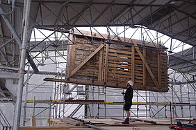 13.6.18 Über eine temporäre Öffnung des Schutzdaches werden die Obergeschosswände mit Hilfe eines Schwerlastkranes versetzt. Hier die Außenwand des Ostgiebels. Fotoaufnahme der Versetzung einer Obergeschosswand mit Hilfe eines Schwerlastkrans. Der Kran hebt die verschalte Wand durch eine temporäre Öffnung des Schutzdaches. An die Unterseite des Bauteils ist ein Seil gebunden, mit dem ein Arbeiter im Inneren die Wand durch die Öffnung führt.
