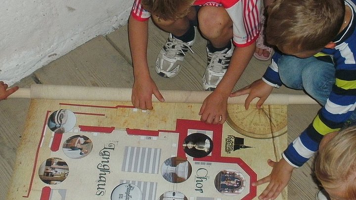 FFM_Schatzsuche_im_Kirchenmuseum Fotoaufnahme der Schatzsuche in der Spitalkirche in Bad Windsheim. Die aufgerollte Schatzkatze ist auf den Holzdielen des Bodens ausgebreitet. Die Kinder sitzen bzw. stehen um die Karte und studieren sie.