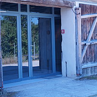 FFM_Archaeologisches_Dorf_Virnsberg_Eingang Fotoaufnahme des Eingangs vom Archäologiemuseum, welches sich in der Schafscheune aus Virnsberg befindet. Das zweiflüglige Holztor ist nach außen geöffnet, die beiden modernen Glastüren dahinter sind geschlossen. Neben der rechten Tür an der Wand befindet sich ein Handfeuermelder.