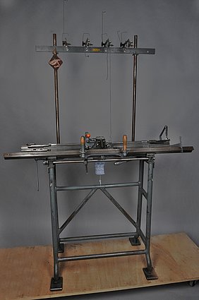 Die Maschine ist groß. Sie hat einen Arbeitstisch. Dort hängt ein Teil einer Socke. Die Socke wird mit der Maschine hergestellt.
