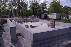 Foto der Betonbodenplatte mit Aufkantung Fotoaufnahme der Betonbodenplatte mit umlaufender Aufkantung. Auf der Platte steht Werkzeug und drei Männer arbeiten darauf. In der linken Bildhälfte steht eine weitere Betonmauer. Im Hintergrund befinden sich vor einer Baumreihe ein Kran, Baumaterial, Container und ein Bauwagen.