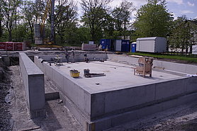 Fotoaufnahme der Betonbodenplatte mit umlaufender Aufkantung. Auf der Platte steht Werkzeug und drei Männer arbeiten darauf. In der linken Bildhälfte steht eine weitere Betonmauer. Im Hintergrund befinden sich vor einer Baumreihe ein Kran, Baumaterial, Container und ein Bauwagen. 