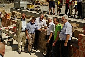 Grundsteinlegung Fotoaufnahme der Grundsteinlegung in der zukünftigen Schürkammer des Badhauses. Inmitten einer Ziegelmauer befindet sich ein Stück gemauerter Sandstein. Sechs Männer haben sich links und rechts davon für ein Foto positioniert. Außerhalb der Baugrube stehen weitere Personen.