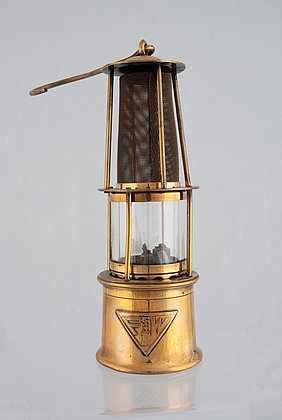 Die Lampe ist aus Messing. Sie ist goldfarben. In der Mitte ist ein Glasgehäuse. Darüber ist ein rundes Metallgitter. Die Lampe hat einen Griff.