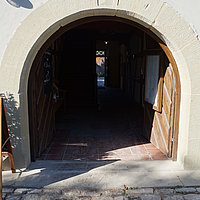 FFM_Gasthaus_zur_Krone_Hintereingang Fotoaufnahme des Hintereingangs vom Gasthaus zur Krone am aktuellen Standort. Die zweiflüglige Holztür ist nach innen geöffnet. Darüber hängt ein Schild, auf dem „Gasthaus zur Krone“ steht. Der Boden vor dem Gebäude ist gepflastert, links ist ein Teil einer Menütafel erkennbar.