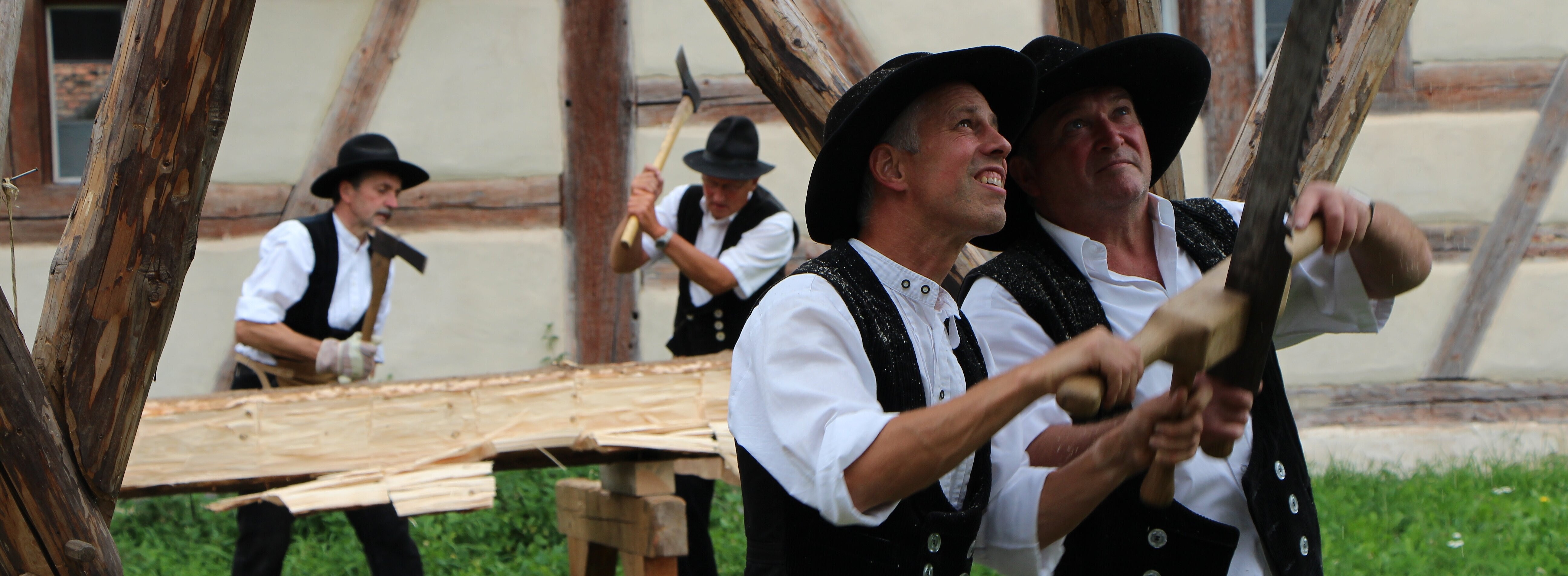 FFM_Zimmerer_beim_Balken_aufsägen Fotoaufnahme von zwei Zimmerern in Zunftkleidung, die mit einer Zweimann-Handsäge an einem Balken im Dachstuhl sägen. Im Hintergrund sind zwei weitere traditionell gekleidete Zimmerer bei der Arbeit.
