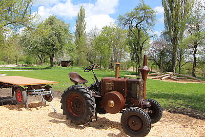 FFM_Erlebnisplatz_Traktor Fotoaufnahme des Traktors auf dem Erlebnisplatz in der Baugruppe Süd. In der Mitte des Bildes steht der alte Traktor, dahinter ein Wagen ohne Bordwände. Dieser wurde allerdings nicht an den Schlepper angehängt.