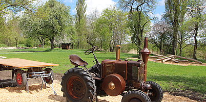 Fotoaufnahme des Traktors auf dem Erlebnisplatz in der Baugruppe Süd. In der Mitte des Bildes steht der alte Traktor, dahinter ein Wagen ohne Bordwände. Dieser wurde allerdings nicht an den Schlepper angehängt.