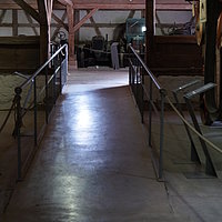 FFM_West_Weiltingen_Rampe Fotoaufnahme der langgezogenen Rampe mit Geländer im Innenraum von der Schafscheuer aus Weiltingen. Hinter der Rampe sind die Fachwerk-Fassade, sowie der offene Dachstuhl erkennbar. In dem Raum werden verschiedene Maschinen ausgestellt.