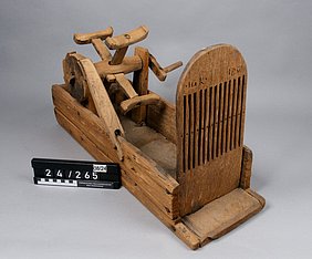 Der Webstuhl ist ein offener Kasten. Hinten ist darauf ein Rad. Es ähnelt einem Mühlrad. Es hat eine Kurbel. Vorne ist eine Holzplatte. Sie ist mit langen Schlitzen versehen. Sie sehen aus wie ein Kamm.