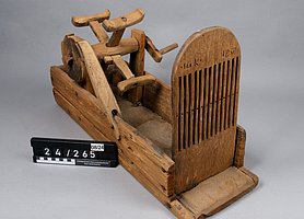 Der Webstuhl ist ein offener Kasten. Hinten ist darauf ein Rad. Es ähnelt einem Mühlrad. Es hat eine Kurbel. Vorne ist eine Holzplatte. Sie ist mit langen Schlitzen versehen. Sie sehen aus wie ein Kamm.
