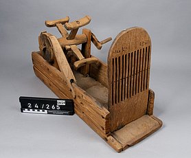 Der Webstuhl ist ein offener Kasten. Hinten ist darauf ein Rad. Es ähnelt einem Mühlrad. Es hat eine Kurbel. Vorne ist eine Holzplatte. Sie ist mit langen Schlitzen versehen. Sie sehen aus wie ein Kamm.