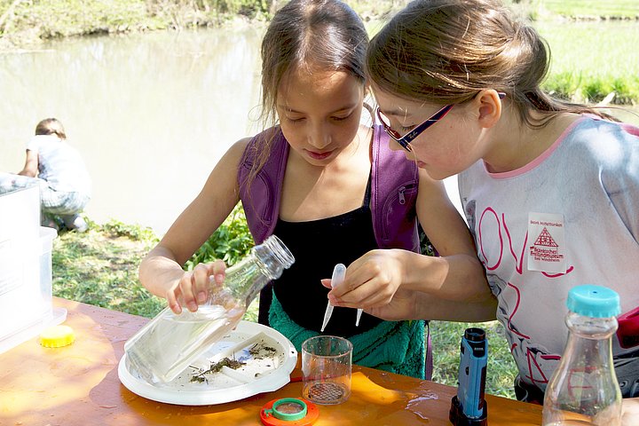 FFM_Wasser_mit_Wasserlabor Fotoaufnahme der Führung „Wasser mit Wasserlabor“. Zwei Mädchen stehen hinter einem Tisch, auf dem sich ein Plastikteller voll Seewasser und eine Becherlupe befinden. Das linke Mädchen tropft mit einer Pipette Wasser ins Becherglas. Sie hält eine Wasserfalsche in der Hand. Dahinter ist der Weiher.