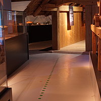 FFM_Archaeologisches_Dorf_Virnsberg_Ausstellung Fotoaufnahme der Ausstellung des Archäologiemuseums, welches sich in der Schafscheune aus Virnsberg befindet. Ein Leitsystem auf dem Boden führt durch die Ausstellung, links und rechts davon stehen Vitrinen mit Objekten.