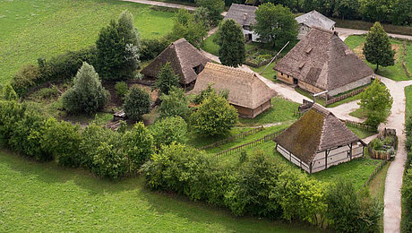 FFM_Mittelalter Fotoaufnahme eines Luftbildes der Baugruppe Mittealter. Die Gruppe von Häusern auf einer Wiese ist von einer Hecke aus Bäumen umgeben und über Wege zugänglich. Umliegend ist die Wiese erkennbar.