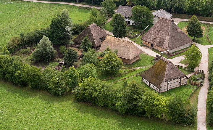 Fotoaufnahme eines Luftbildes der Baugruppe Mittealter. Die Gruppe von Häusern auf einer Wiese ist von einer Hecke aus Bäumen umgeben und über Wege zugänglich. Umliegend ist die Wiese erkennbar. 