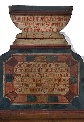 Das Kopfteil des Betts ist bunt bemalt. Es ist blau, rot und beige. Darauf steht der Name der Braut. Sie heißt Maria Elisabetha Reichert. Auch steht eine Jahreszahl darauf. Das Bett wurde 1836 gebaut.