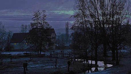 FFM_Nachtwächter Fotoaufnahme vom Freilandmuseum bei Nacht. Im Vordergrund befindet sich das schneebedeckte Ufer eines kleinen Baches. Links dahinter ist eine eingezäunte Weide, rechts stehen kahle Bäume. Im Hintergrund ist das Köblerhaus aus Schwimbach erkennbar.