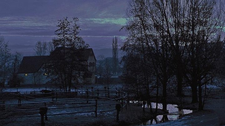 FFM_Nachtwächter Fotoaufnahme vom Freilandmuseum bei Nacht. Im Vordergrund befindet sich das schneebedeckte Ufer eines kleinen Baches. Links dahinter ist eine eingezäunte Weide, rechts stehen kahle Bäume. Im Hintergrund ist das Köblerhaus aus Schwimbach erkennbar.