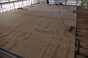 6.6.18 Nur für kurze Zeit war die Erdgeschossbalkenlage vollständig zu sehen, bevor sie mit Schaltafeln als Arbeitsebene für den weiteren Aufbau des Obergeschosses belegt wurde. Fotoaufnahme der hölzernen Schaltafeln, die als Arbeitsebene für den weiteren Aufbau des Obergeschosses auf der Erdgeschossbalkenlage ausgelegt wurden. Die Schaltafeln bedecken den Großteil des Bodens, lediglich am rechten Rand sind die Holzbalken noch erkennbar.